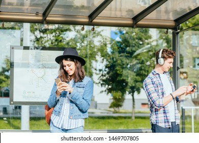 Youth teenagers people waiting for transport on tram or bus stop. Brunette caucasian woman and hipster student man holds smartphones on hands. - Powered by Shutterstock