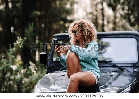 Youthful adult woman sitting on the car front and using mobile phone with forest green woods around. People enjoying travel and vehicle in the nature outdoors. Vehicle traveler female using connection