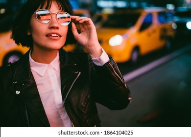 Youthful Female Party Girl In Optical Glasses With Neon Lights Reflection Looking Up During Evening Walk On Manhattan, Millennial Teenager In Spectacles Standing At Urban Street During Nightlife