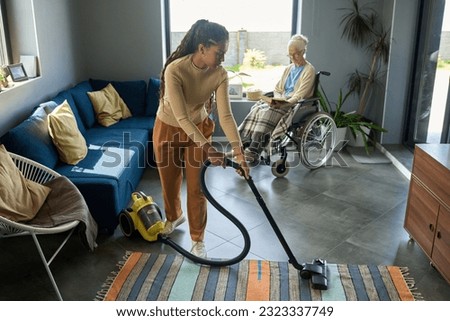 Youthful girl with vacuum cleaner helping her grandmother with disability