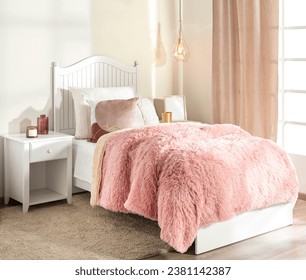 A youthful girl's bedroom showcasing a soft pink fur blanket on a white bed frame, decorative pillows, a white night table with a drawer, an elegant hanging bulb light, cream curtains, sunlit ambiance
