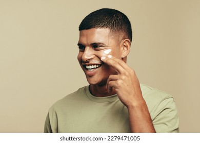 Youthful man smiles with joy as he introduces a beauty cream to his skincare routine. Happy young man pampering his face with a moisturizing product to achieve a glowing and radiant skin tone.
