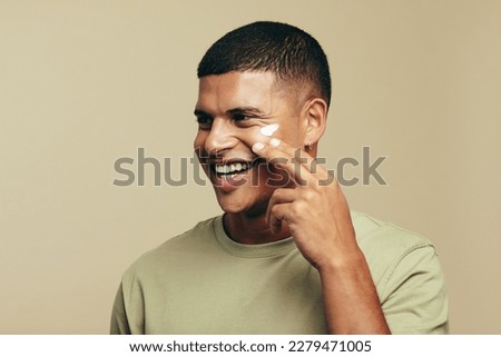 Youthful man smiles with joy as he introduces a beauty cream to his skincare routine. Happy young man pampering his face with a moisturizing product to achieve a glowing and radiant skin tone.