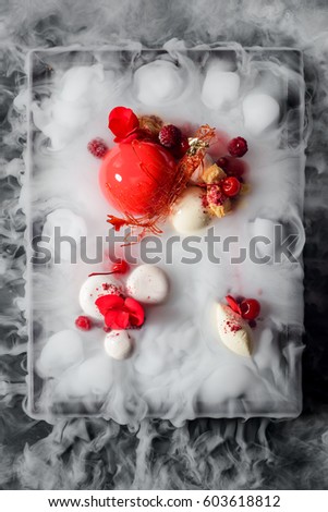 Yummy mousse cake, yogurt spheres, cherries, raspberries, a caramel decoration and fresh flowers in dry ice mist, a top-view image. Molecular food.