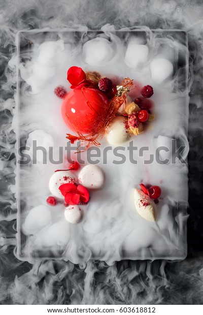 Yummy mousse cake, yogurt spheres,\
cherries, raspberries, a caramel decoration and fresh flowers in\
dry ice mist, a top-view image. Molecular\
food.
