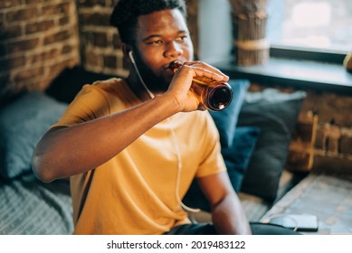 Yung Attractive Black Man With Dark Skin Sitting At Home Indoors And Drinking A Bottle Of Beer. Moody Lifestyle Image Authentic Portrait