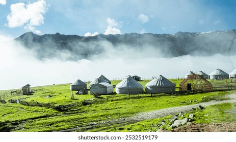 Yurts dot a vibrant green meadow, enclosed by rustic wooden fences, beneath a misty sky with mountains softly silhouetted in the background. - Powered by Shutterstock