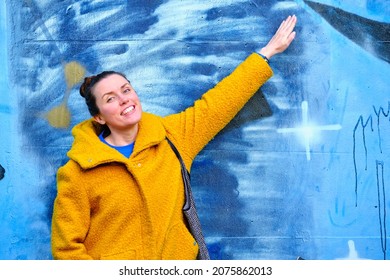 Zagreb, Croatia- 10 10 2021: Beautiful Young Caucasian Girl With Bun And Yellow Coat Showing Amazing Blue Graffitti Artwork