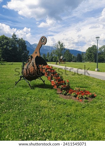 In Zakopane, a large wooden vase, a step full of small vases with colorful, red and orange flowers, on green grass. on the right, a post, in the background trees, mountains, blue sky and white clouds.
