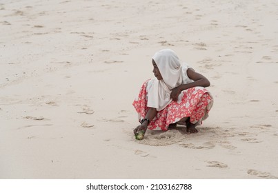 Zanzibar, Tanzania -12.01.2021: Young local girl playing in the sand on Nakumba beach. Moslem girl enjoying the sunny weather on the beach. Child alone on the beach.