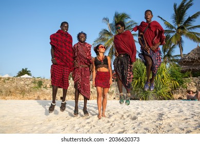 Zanzibar, Tanzania, January 27, 2021: Four masses in traditional red clothes and European girl jumping on the beach, in the background of palm trees and roof bungalows