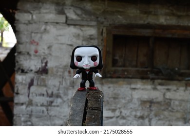 Zaporizhzhia, Ukraine - July 3, 2022: Illustrative Editorial Of Funko Pop Action Figure Of Billy The Puppet From Popular Horror Movie Saw. Toy Standing On Wooden Wheel In Deserted Yard Near Lost Shed.