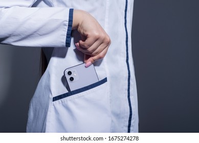 Zaporozhye. Ukraine. February 2020. Studio Portrait Of A Girl In A White Coat. The Doctor Holds In His Hand An IPhone 11 From Epl. On A Gray Background.