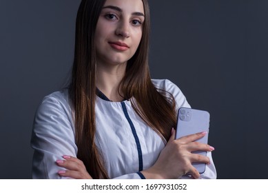 Zaporozhye. Ukraine. February 2020. Studio Portrait Of A Girl In A White Coat. The Doctor Holds In His Hand An IPhone 11 From Epl. On A Gray Background.