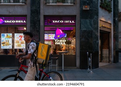 Zaragoza, Spain - August 23, 2022: A Glovo Delivery Man On A Bike Rides Past A Taco Bell Fast Food Restaurant, Where Tacos, Burritos And Mexican Food Are Sold, In The City Of Zaragoza