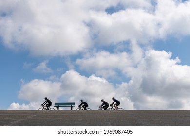 Zeeland, The Netherlands-May 2021: Low Angle View Of An Empty Park Bench With A Group Of Cyclists Passing By On Top Of One Of The Asphalt Covered Dikes Against A White Clouded Blue Sky