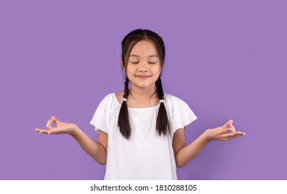 Zen. Calm Asian Kid Girl Meditating Standing Over Purple Background With Eyes Closed. Studio Shot