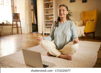 Zen, peace, balance, concentration and technology concept. Attractive senior woman sitting on carpet in front of open laptop keeping eyes closed and legs crossed, meditating to nature sounds