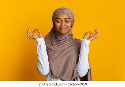 Zen. Pleased Black Muslim Woman In Headscarf Meditating With Closed Eyes, Making Mudra Gesture With Fingers, Standing Over Yellow Background