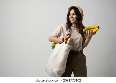 Zero waste concept. Young brunette woman holding cotton eco bag with organic fruits and vegetables on a shoulder and holding bananas in a hand. Using reusable crochet net bag for grocery shopping.