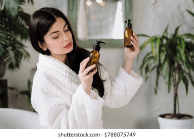 Zero Waste Lifestyle Concept. Minded Young Adult Woman In Bathrobe, Spending Morning At Bathroom, Choosing Personal Hygiene Items, Holding Glass Bottle With Cosmetic Care Product