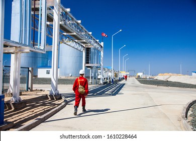 Zhanazhol, Aktobe Region, Kazakhstan, May 04 2012: Oil Refinery Plant In Desert. Worker In Red Work Wear On Pipes And Oil Storage Tanks And Blue Sky Background. Red Weather Vane. CNPC-AMG Company