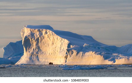 A zodiac full of tourist viewed against a large iceberg with the light of the midnight sun on the summer solstice, Cierva Cove, Antarctica