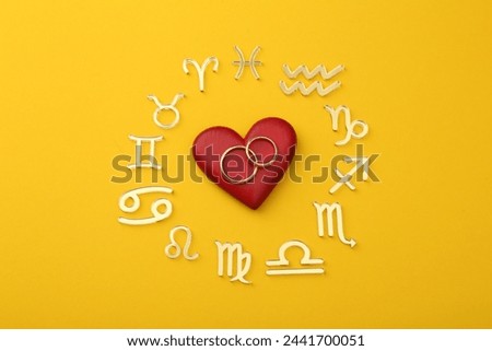 Zodiac signs, red heart and wedding rings on yellow background, flat lay