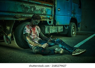 Zombie Apocalypse. A Scary Bloodied Zombie Sits On The Ground Leaning On An Old Car In A Ruined City. Halloween. Horror Movie. 