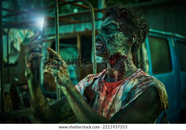 Zombie apocalypse. A scary bloodied zombie stands by an\
old car in a ruined city and uses a smartphone. Halloween. Horror\
movie. 