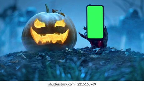 Zombie hand holds smartphone with green screen out of grave near Glowing Halloween pumpkin in the cemetery. Holiday event halloween concept. - Shutterstock ID 2215163769