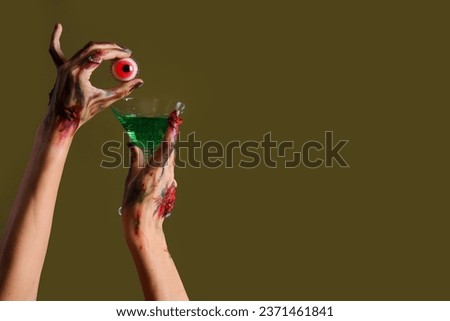 Zombie hands holding glass with cocktail and eyeball on green background. Halloween celebration