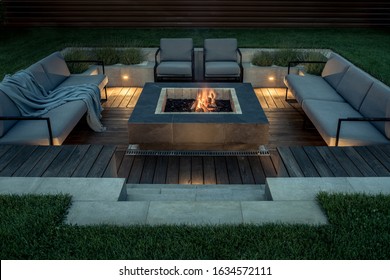 Zone for relax with a wooden floor and a tiled stair outdoors. There is a burning fire pit, gray sofas and armchairs, plaid, luminous lamps. Horizontal. - Shutterstock ID 1634572111