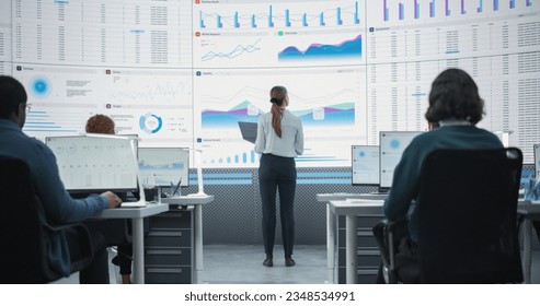 Zoom Out Back View Shot Of Female Data Scientist Using Laptop And Analyzing Information On Big Digital Screen in Monitoring Office. Diverse Employees Working On Desktop Computers For Big Data Company