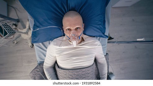 Zoom In View Of Bald Man With Respirator Looking At Camera While Lying On Bed Near Life Support Machine In Oncology Clinic