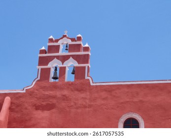 Zoomed view of a red colonial church bell tower with white accents and visible bells, contrasted against a clean blue sky in a traditional Mexican town in Campeche.
