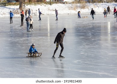 ZUTPHEN, NETHERLANDS - Feb 12, 2021: Scenery With Ice Skating Father Pulling A Sledge With Child Lit Up By Sunset Light On Open Water Frozen Canal With Others Riding Behind