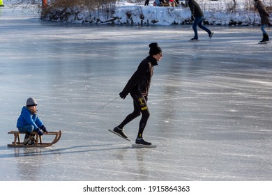 Zutphen, The Netherlands - February 12, 2021: Ice Skating Father Pulling A Sledge With Child Lit Up By Sunset Light On Open Water Frozen Canal With Others Riding Behind