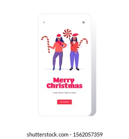 african american couple in santa claus hats holding sweetmeats candy cane and lollipop merry christmas happy new year winter holidays celebration concept smartphone screen online mobile app full