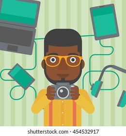 An african-american man taking photo with digital camera. Man surrounded with gadgets. Man using many electronic gadgets. Vector flat design illustration. Square layout.
