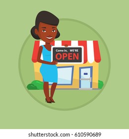 African-american shop owner holding open signboard. Shop owner standing in front of small store. Woman inviting to come in shop. Vector flat design illustration in the circle isolated on background.