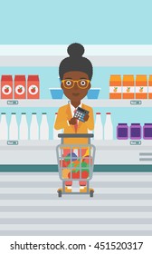 An african-american woman at the supermarket with calculator and supermarket trolley full with products. Woman checking prices with calculator. Vector flat design illustration. Vertical layout.