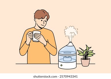 Air humidifier on table near man to avoid dryness in room and create comfortable atmosphere. Guy smiles and drinks coffee while looking at working air humidifier with steam escaping