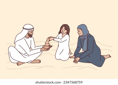 Arab family sits in desert with dunes, playing with daughter with sand and enjoying joint weekend. Happy family from arab emirates or qatar dressed in ethnic clothes, for concept oriental culture