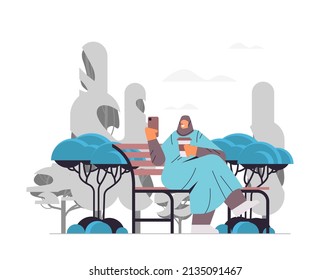 arab woman drinking coffee and using smartphone in urban park girl sitting on wooden bench landscape background