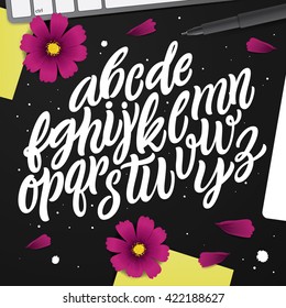 Artwork with ABC set. Black desk with vector characters, post-it note, pen and pink flowers. Typography Background. Cursive Modern Lettering Alphabet. Perfect font for your projects.