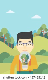 An asian man standing on the background of green forest and holding in hands a big lightbulb with trees inside. Green energy concept. Vector flat design illustration. Vertical layout.