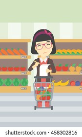 An asian thoughtful woman standing at the supermarket with supermarket trolley full with products and holding a shopping list in hands. Vector flat design illustration. Vertical layout.