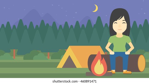 An asian woman sitting on a log near a fire on a background of camping site with tent. Woman sitting near a campfire at a campsite. Vector flat design illustration. Horizontal layout.