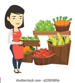 Asian Worker Of Grocery Store Standing In Front Of Section With Vegetables And Fruits. Worker Of Grocery Store Holding A Box With Apples. Vector Flat Design Illustration Isolated On White Background.